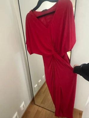 Röd långklänning med knutdetalj - Säljer en elegant röd långklänning med v-ringning och snygg knutdetalj framtill. Klänningen har lös passform upptill, korta ärmar och ett mjukt, följsamt tyg som ger ett fint fall. Perfekt för dig som vill sticka ut med en färgstark look.