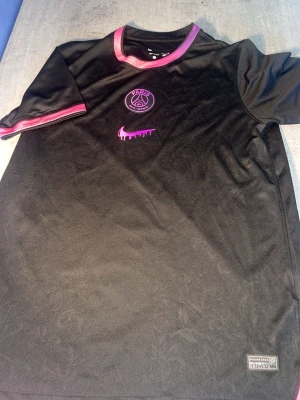 Svart PSG x Nike t-shirt - Snygg svart t-shirt från Nike i samarbete med Paris Saint-Germain. Tröjan har rosa detaljer på krage och ärmslut, samt PSG-logga och en unik rosa Nike swoosh på bröstet. Tillverkad i ett lätt och ventilerande material, perfekt för träning eller streetwear.