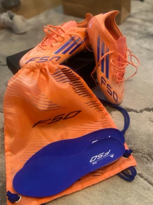 Adidas F50 orange fotbollsskor - Säljer ett par Adidas F50 fotbollsskor i en grymt snygg orange färg med blå detaljer och klassiska tre ränder på sidan. Skorna har snörning, låg profil och kommer med matchande påse och blå innersulor. Perfekta för snabba spelare som vill sticka ut på planen.
