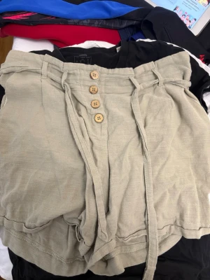 Grön  linneshorts med knappar och knyt - Snygga gröna shorts i linne med hög midja, dekorativa träknappar framtill och ett knytband i midjan. Shortsen har avslappnad passform och fickor både fram och bak. Perfekta för varma dagar och enkel att styla.