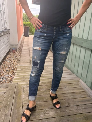 Blå lågmidjade slitna jeans - Säljer ett par blå  jeans med slitningar och patchdetaljer. Jeansen har uppvikta benslut. Perfekta för en cool och avslappnad stil. Materialet är klassisk denim i bomull.