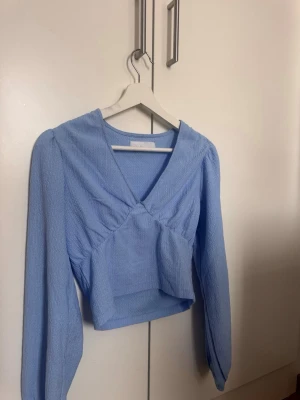 Ljusblå croppad topp Samsa Collection - Trendig ljusblå croppad topp från Samsa Collection med v-ringning och långa ärmar. Toppen har en snygg omlott-look framtill och är tillverkad i ett strukturerat tyg som ger en cool känsla. Perfekt att styla med höga jeans eller kjol.