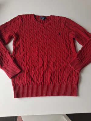Röd kabelstickad tröja Polo Ralph Lauren - Snygg röd kabelstickad tröja från Polo Ralph Lauren med rund halsringning och klassisk marinblå logga broderad på bröstet. Tröjan har långa ärmar och ribbade muddar vid ärmslut och nederkant. Perfekt för dig som gillar tidlös stil. Varan är i mycket bra skick i storlek S. Hör av dig vid frågor