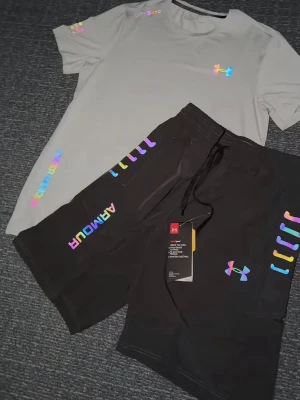Grå träningsset från Under Armour - Snyggt set från Under Armour med en ljusgrå t-shirt och svarta shorts. Båda plaggen har regnbågsfärgade loggor och detaljer, samt tryckt text på ärm och ben. Perfekt för dig som gillar sportig och färgglad stil. Storlek S, helt ny 