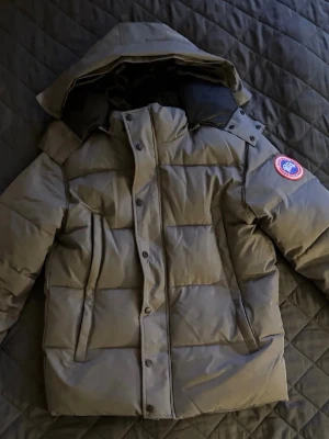  Canada Goose jacka  - Säljer en grå dunjacka från Canada Goose med huva. Jackan har stora, praktiska fickor framtill och stängs med både dragkedja och knappar. 