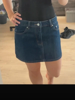 Mörkblå jeanskjol - Säljer en klassisk mörkblå jeanskjol i kort modell med fem fickor och knappstängning framtill. Kjolen har en enkel och tidlös design som passar till det mesta. Perfekt för dig som gillar denim och vill ha en basic kjol i garderoben.