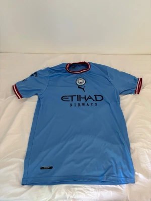 Manchester City matchtröja Puma - Ljusblå Manchester City matchtröja från Puma med Etihad Airways-tryck på bröstet och Haaland 9 på ryggen. Tröjan har korta ärmar, klubbmärke på bröstet och röda detaljer vid krage och ärmslut. Perfekt för fotbollsfans!