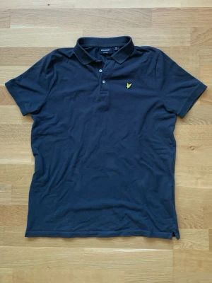 mörkgrå pikétröja från Lyle & Scott - Klassisk mörkgrå pikétröja från Lyle & Scott med gul broderad logga på bröstet. Tröjan har krage, korta ärmar och knappar framtill. Perfekt till jeans eller shorts för en clean och stilren look.