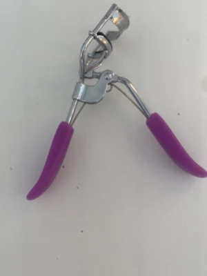 Ögonfransböjare med lila handtag - En klassisk ögonfransböjare i metall med ergonomiska handtag i lila gummi för extra grepp. Böjaren har en silvrig finish och en mjuk kudde för att skydda fransarna. Perfekt för att skapa snygg böj på fransarna snabbt och enkelt.