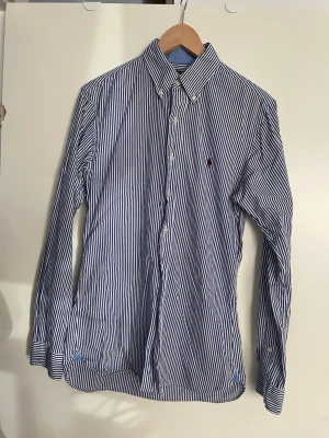 Randig skjorta från Ralph Lauren SP slim fit - Klassisk blå- och vitrandig skjorta från Ralph Lauren med broderad logga på bröstet. Skjortan har lång ärm, knappar framtill och krage. Tillverkad i bomull för en skön och luftig känsla. Perfekt för en stilren och tidlös look.