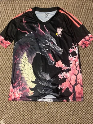 Japan JFA tröja med drakmotiv - Unik svart sporttröja från Japan JFA med stort drakmotiv, körsbärsblommor och japanskt tempel. Tröjan har korta ärmar med rosa detaljer och tryck, samt rund halsringning. Tillverkad i ett lätt och snabbtorkande material, perfekt för träning eller match.