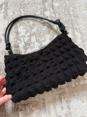 Svart axelväska med textur - Trendig svart handväska med unik textur och vågig kant. Väskan har ett kort axelband i svart konstläder med knutdetalj. Perfekt för dig som gillar statement-accessoarer och vill sticka ut lite extra.