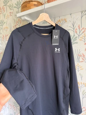 Svart långärmad tröja Under Armour - Snygg svart långärmad träningströja från Under Armour med rund hals och diskret logga på bröstet. Tillverkad i ett stretchigt funktionsmaterial som andas, perfekt för träning eller löpning. Snygga sömdetaljer ger en sportig look.