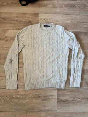 Beige kabelstickad tröja Dressmann - Kabelstickad tröja från Dressmann i beige Tröjan har rund halsringning, ribbade muddar och nederkant samt ett klassiskt flätmönster. Perfekt för dig som gillar en enkel men snygg stil. De ändå defekten är en blyech fläck på armen som ni ser på bild 1 annars är den som nu använd fåtals gånger 