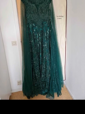 Grön långklänning med paljetter - Elegant grön långklänning med glittriga paljettdetaljer och transparent tyll över axlarna. Klänningen har en figurnära överdel och faller vackert mot golvet. Perfekt för speciella tillfällen där du vill sticka ut.