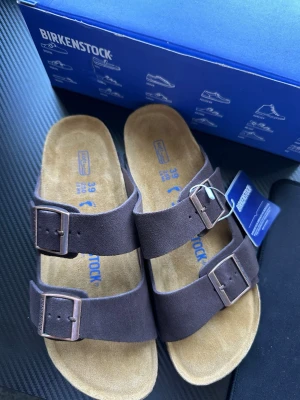 Bruna Birkenstock Arizona sandaler - Säljer helt nya par klassiska Birkenstock Arizona sandaler i mörkbrunt mocka med två breda remmar och justerbara metallspännen. Fotbädden är i mjukt naturfärgat material och sulan är i kork och gummi för extra komfort. Perfekta för en avslappnad och stilren look.