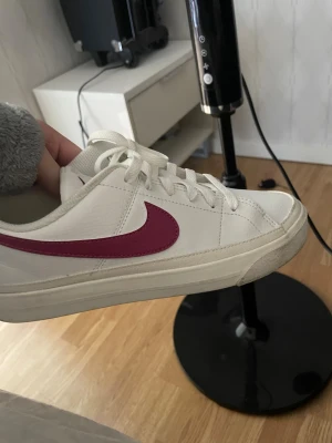 Vita Nike sneakers med rödaktig swoosh - Säljer ett par klassiska vita Nike sneakers med rödaktig swoosh på sidan. Skorna har lågt skaft och vita snören. Perfekta för dig som gillar en clean och sportig stil med en färgklick.