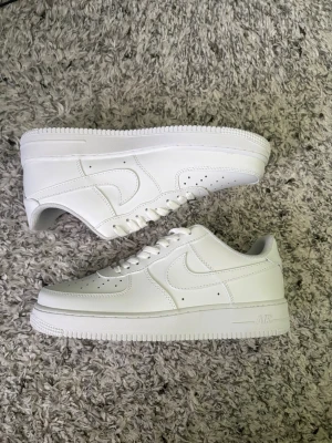 Nike Air Force 1 vita sneakers - Klassiska helvita Nike Air Force 1 sneakers i lågt utförande. Skorna är tillverkade i slätt läder med perforeringar på tån för ventilation och har den ikoniska swoosh-loggan på sidan. Tjock vit sula med präglad 'AF1'-detalj och snörning framtill.