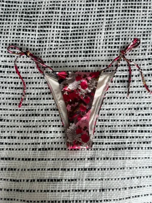 Blommig bikiniunderdel med knyt - Säljer en snygg bikiniunderdel från Urban Outfitters i strl XS med blommigt mönster i rött och rosa. Modellen har smala knytband i sidorna för justerbar passform och en klassisk trekantsform. Orginalpris är 239kr, den är helt oanvänd med lappen kvar samt hygien klistermärket.