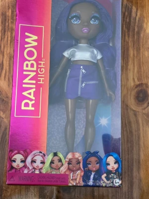 Rainbow High ,Krystal Bailey - Säljer en Rainbow High docka i originalförpackning. Dockan har lila hår, vit kort topp och lila kjol med dragkedjedetaljer. Förpackningen är färgglad med illustrationer av andra karaktärer från serien. Perfekt för samlare eller som present.