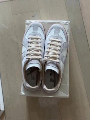 Maison Margiela skor 👟  - Snygga sneakers i vitt och beige med detaljer i mocka och slät läder. Skorna har klassisk låg profil, vita snören och en brun gummisula som ger retrovibbar. Perfekta för dig som gillar stilrena och tidlösa sneakers.