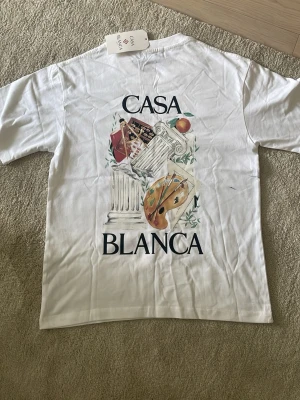 Vit t-shirt från Casablanca - Vit t-shirt från Casablanca med konstnärligt tryck av pelare, palett och frukt på både bröstet och ryggen. Klassisk rund hals och korta ärmar. Stora trycket på ryggen och mindre på framsidan ger en unik look. Perfekt för dig som gillar stilrena men kreativa plagg.Helt ny.