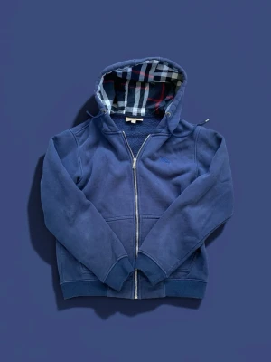 Blå hoodie med dragkedja och rutigt foder - Snygg blå hoodie från Burberry med dragkedja och klassiskt rutigt foder i huvan och längs insidan. Hoodien har ribbade muddar, fickor framtill och är gjord i mjukt bomullsmaterial. Perfekt för en chill och stilren look.