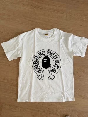 Vit BAPE x Chrome Hearts t-shirt - Säljer en vit t-shirt från BAPE med stor Chrome Hearts-inspirerad print i svart på bröstet. Klassisk rund hals och korta ärmar. Materialet är mjuk bomull och passformen är normal. Perfekt för dig som gillar streetwear och unika samarbeten.