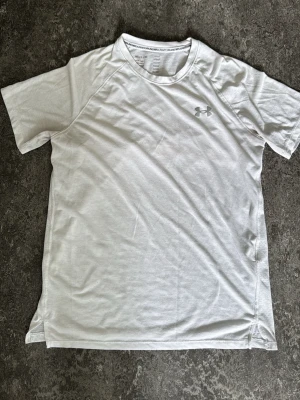 Vit tränings-tshirt från Under Armour - Snygg vit tränings-tshirt från Under Armour med diskret logga på bröstet. Tillverkad i ett lätt och snabbtorkande funktionsmaterial som passar perfekt till gymmet eller löprundan. Klassisk passform med kort ärm och rund hals.