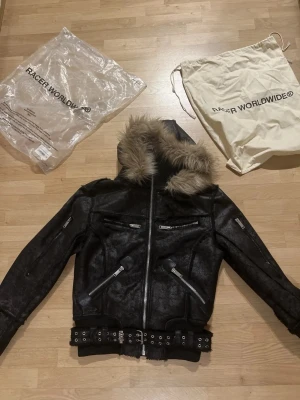 Racer Worldwide skinnjacka - Snygg svart skinnjacka från Racer Worldwide med avtagbar huva och fuskpäls. Jackan har flera dragkedjeförsedda fickor, bälte i midjan och coola detaljer som ger en edgy look. Perfekt för dig som vill sticka ut och hålla värmen. Använd en gång. Passa på.