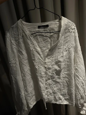 Vit blommig blus från Gina Tricot - Söt vit blus från Gina Tricot med broderade blommor och v-ringning. Blusen har långa ärmar och fina spetsdetaljer längs halsringningen. 