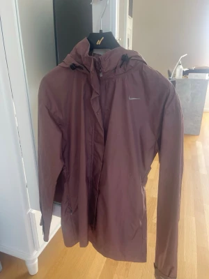 Brun vindjacka från Nike med huva - Snygg brun vindjacka från Nike med justerbar huva och dragkedja framtill. Jackan har en minimalistisk design med diskret Nike-logga på bröstet och reflexdetalj på ryggen. Tillverkad i lätt och vindtätt material, perfekt för blåsiga dagar.