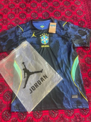 Brasil x Jordan matchtröja svart/blå - Snygg svart och blå matchtröja från Brasilien med Jordan-logga. Tröjan har gula detaljer, CBF-emblem på bröstet och gröna inslag på sidorna. Tillverkad i lätt och ventilerande material, perfekt för fotboll eller träning.