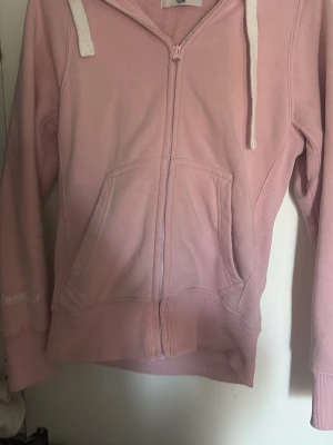 Ljusrosa Russemerch hoodie  - Ljusrosa russemerchhoodie med dragkedja och vita snören i huvan. Hoodien har två stora fickor framtill och ribbade muddar vid ärmar och nederkant. Tillverkad i mjukt bomullsmaterial som känns skönt mot huden. Dragkedje färgen har låssat lite men inget man direkt tänker på 