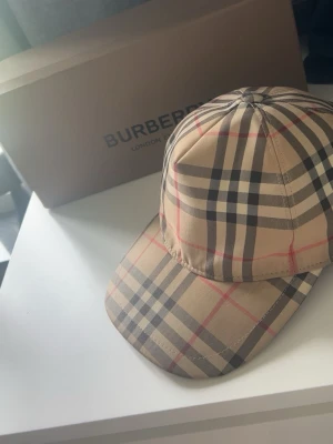 Beige keps från Burberry med rutmönster - Säljer en klassisk beige keps från Burberry med det ikoniska rutmönstret i svart, vitt och rött. Kepsen har böjd skärm och justerbar rem bak. Perfekt accessoar för dig som vill ha en stilren och tidlös look.