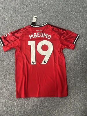 Manchester United matchtröja röd - Säljer en röd Manchester United matchtröja från Adidas med tryck 'MBEUMO 19' på ryggen. Tröjan har klubbmärke, Adidas-logga och Snapdragon sponsortryck på bröstet samt svarta och vita detaljer på ärmarna. Tillverkad i ett lätt och ventilerande material.