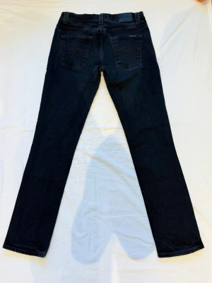 Mörkblå raka jeans från Nudie Jeans - Snygga mörkblå jeans från Nudie Jeans med rak passform och klassisk femficksdesign. Jeansen har subtila sömmar på bakfickorna och är tillverkade i slitstark denim. Perfekta för dig som gillar en clean och tidlös look.