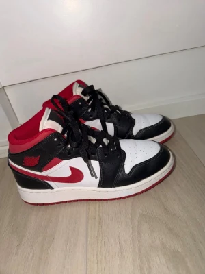 Air Jordan 1 Mid svart/röd/vit - Säljer ett par äkta Nike Air Jordan 1 Mid sneakers i svart, rött och vitt. Storlek 37,5. Klassisk design med svart läderöverdrag, vit bas och röda detaljer som swoosh och foder. Skorna har snörning och den ikoniska Air Jordan-loggan på sidan. Har bara använt dessa skor 1-2 gånger max, så de är i princip oanvända och nya. 