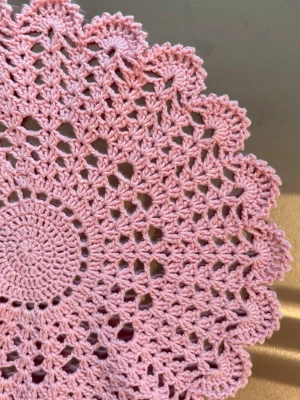 Rosa virkad duk med spetsmönster - Säljer en rund, handvirkad duk i ljusrosa bomullsgarn med detaljerat spetsmönster och vågig kant. Perfekt som dekorativt underlägg på bordet för att ge en personlig touch till din inredning. Kan även göra i andra färger!
