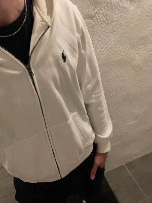 Vit hoodie från Polo Ralph Lauren - Vit hoodie med dragkedja från Polo Ralph Lauren. Klassisk design med huva, två fickor framtill och den ikoniska mörkblå loggan broderad på bröstet. Tillverkad i mjukt bomullsmaterial som är skönt. Finns inga fläckar eller liknande 