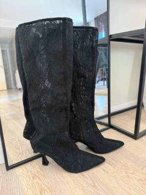 Svarta spetsstövlar med klack - Säljer ett par höga svarta stövlar med spetsdetaljer och blommönster. Stövlarna har spetsig tå och smal klack, vilket ger en elegant och trendig look. Perfekta för dig som vill sticka ut med en unik stil. Materialet är genombruten spets med blommönster.