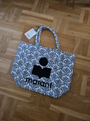 Mönstrad shoppingväska från Isabel Marant - Säljer en stor shoppingväska från Isabel Marant i vit canvas med blått, detaljerat mönster och svart logotyptryck. Väskan har två rejäla handtag och är perfekt för att bära allt du behöver. Snygg och trendig accessoar för dig som gillar statement-plagg.
