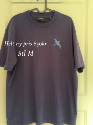 Lila t-shirt med blå fågeltryck - Snygg lila t-shirt från Acne Studios med ett litet blått fågeltryck på bröstet. Klassisk rund halsringning och avslappnad passform. Perfekt för dig som gillar stilrena plagg med en twist.
