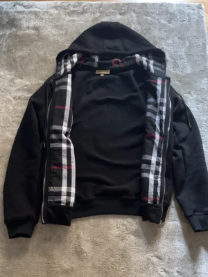 Svart hoodie med dragkedja Burberry - Säljer en svart hoodie från Burberry med dragkedja och klassiskt rutigt mönster i huvan. Hoodien har fickor framtill, ribbade muddar och justerbar dragsko i huvan. Perfekt för dig som gillar stilrena plagg med en twist.