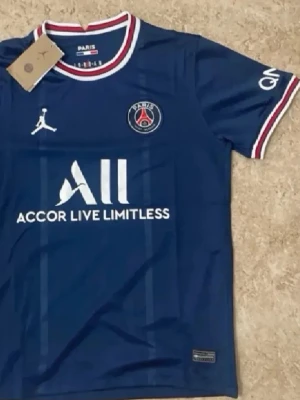 Blå PSG matchtröja från Jordan - Snygg blå PSG matchtröja med vita och röda detaljer på krage och ärmslut. Tröjan har PSG-logga på bröstet, Jordan-logga och sponsortryck framtill. Tillverkad i lätt och ventilerande material, perfekt för fotboll eller träning.