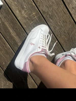 Vita sneakers Polo Ralph Lauren -  vita sneakers från Polo Ralph Lauren med rosa glitterdetaljer vid hälen och rosa broderad logga på sidan. Skorna har klassisk låg profil, vita snören och ovandel i slätt material. Perfekta för dig som vill ha en clean look med lite extra bling.