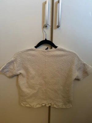 Vit croppad t-shirt i frotté - En vit croppad t-shirt med korta ärmar och rund hals. T-shirten är gjord i ett mjukt frottématerial och har en synlig söm mitt bak samt lätt rynkad nederkant. Perfekt för en avslappnad och trendig look.