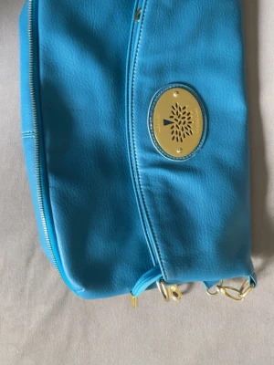 Mulberry med gulddetaljer - Snygg blå kuvertväska replika från Mulberry i konstläder med guldfärgad logoplatta framtill. Väskan har dragkedja och avtagbar axelrem i guld. Perfekt för dig som vill ha en färgklick till outfiten och gillar stilrena detaljer.