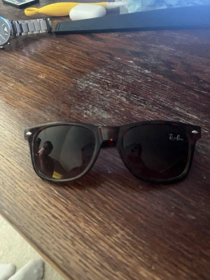 Ray-Ban Wayfarer solglasögon brun - Säljer ett par klassiska Ray-Ban Wayfarer solglasögon i mörkbrun sköldpaddsmönstrad plast. De har svarta glas och den ikoniska Ray-Ban-loggan på linsen. Snygg och tidlös modell som passar till allt.