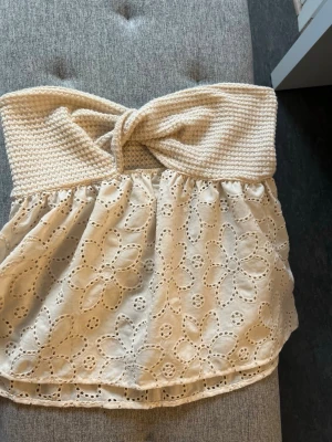 Krämvit axelbandslös spets tubtopp - Söt krämvit  axelbandslös tubtopp med virkad överdel och broderad spets med blommönster. Toppen har en twistad detalj framtill och är perfekt för varma dagar. Materialet känns luftigt och toppen har en romantisk vibe.